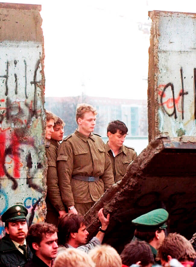 Guardias fronterizos de Alemania del Este miran a través de un agujero en el muro de Berlín después de que los manifestantes derribaran un segmento del muro en la puerta de Brandenburgo el sábado 11 de noviembre de 1989 (Foto © Lionel Cironneau | AP).