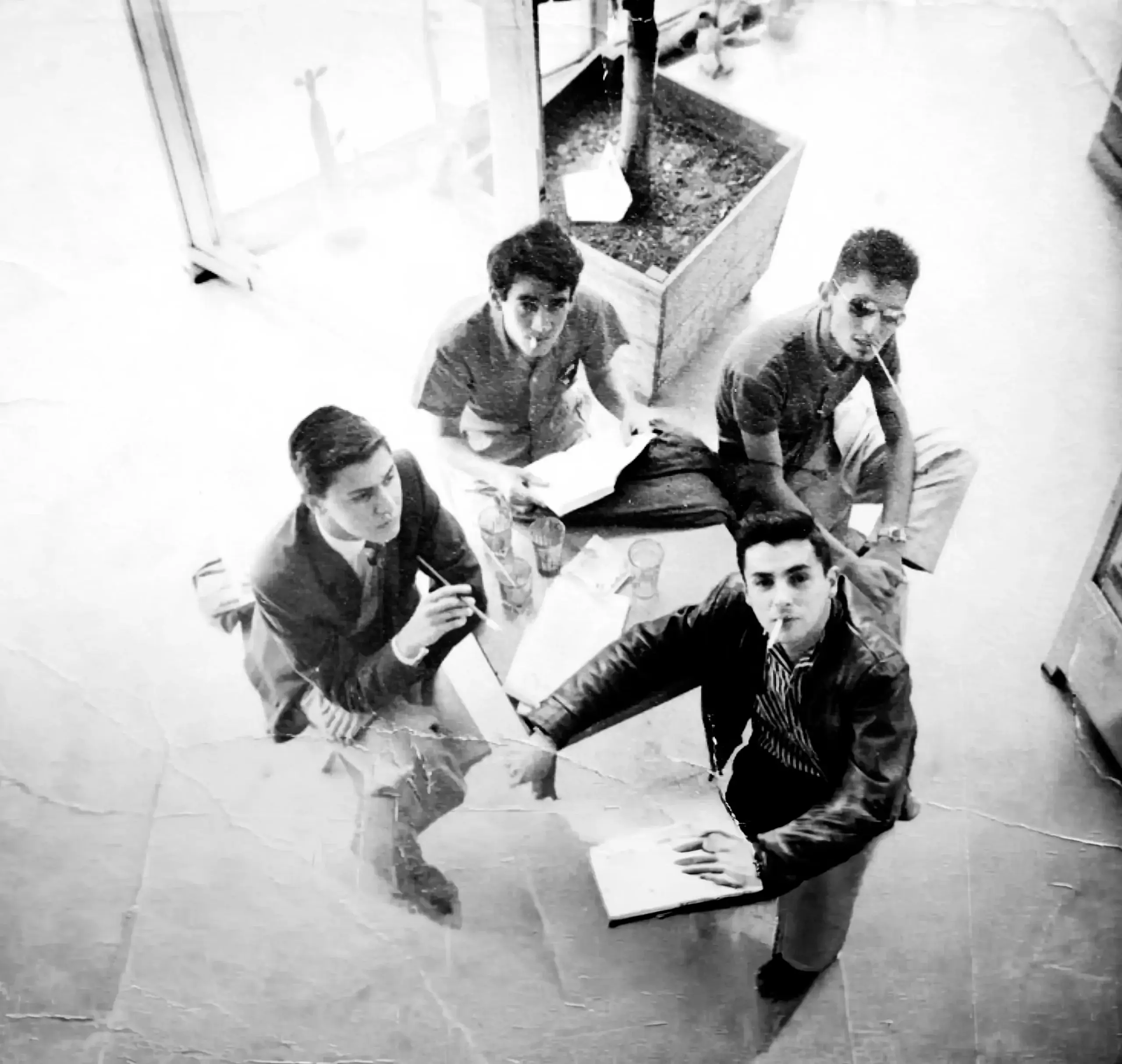 Alberto Escobar, Gonzalo Arango, Alirio Ozogar y Malgrem Restrepo miran hacia la cámara desde el interior de un edificio, fotografiados desde arriba; dos de ellos están fumando y Gonzalo sostiene un libro abierto.