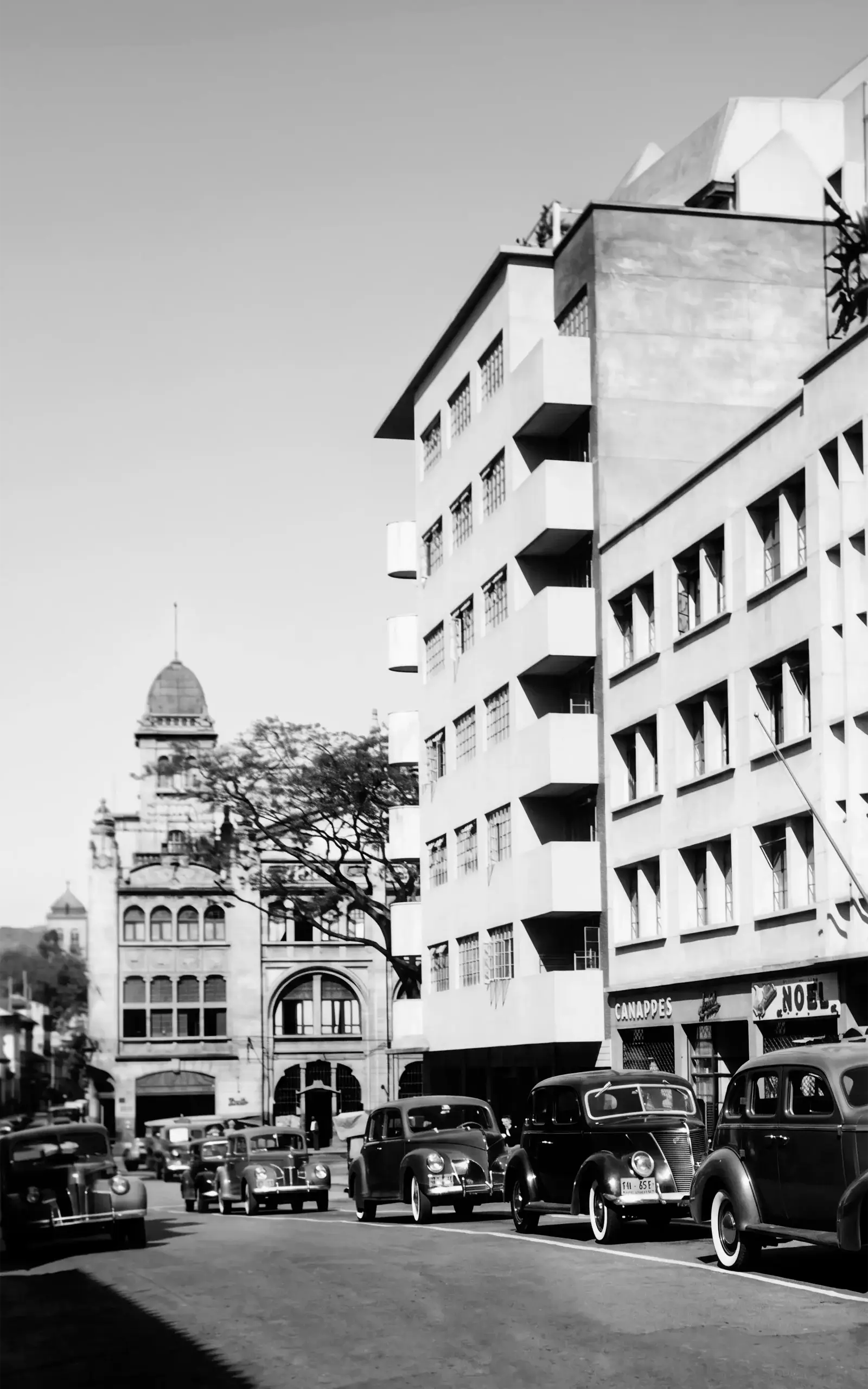 Una calle de Medellín con automóviles antiguos, edificios de varias plantas y una torre con cúpula al fondo.