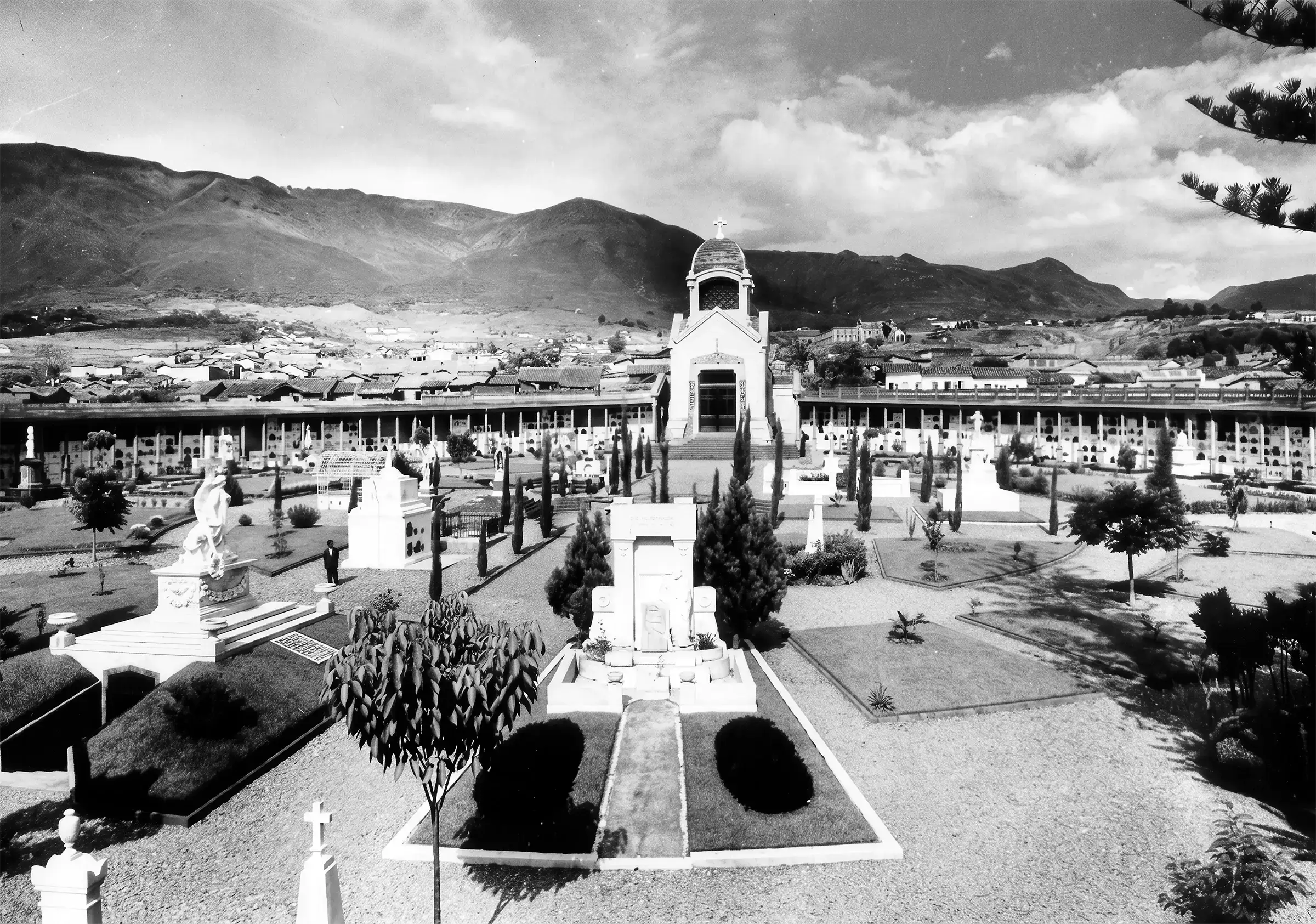 Vista del cementerio de San Pedro con capilla central, esculturas y jardines, rodeado de montañas.