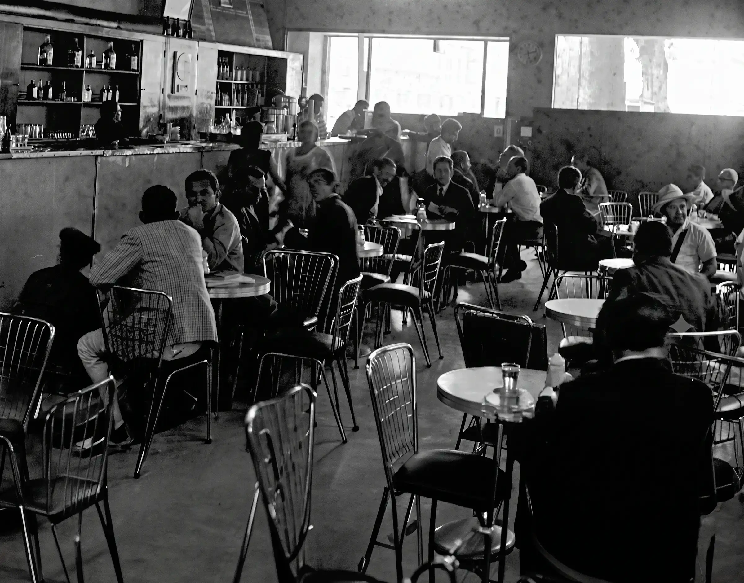 Interior del café La Bastilla con clientes sentados alrededor de mesas pequeñas y una barra al fondo.